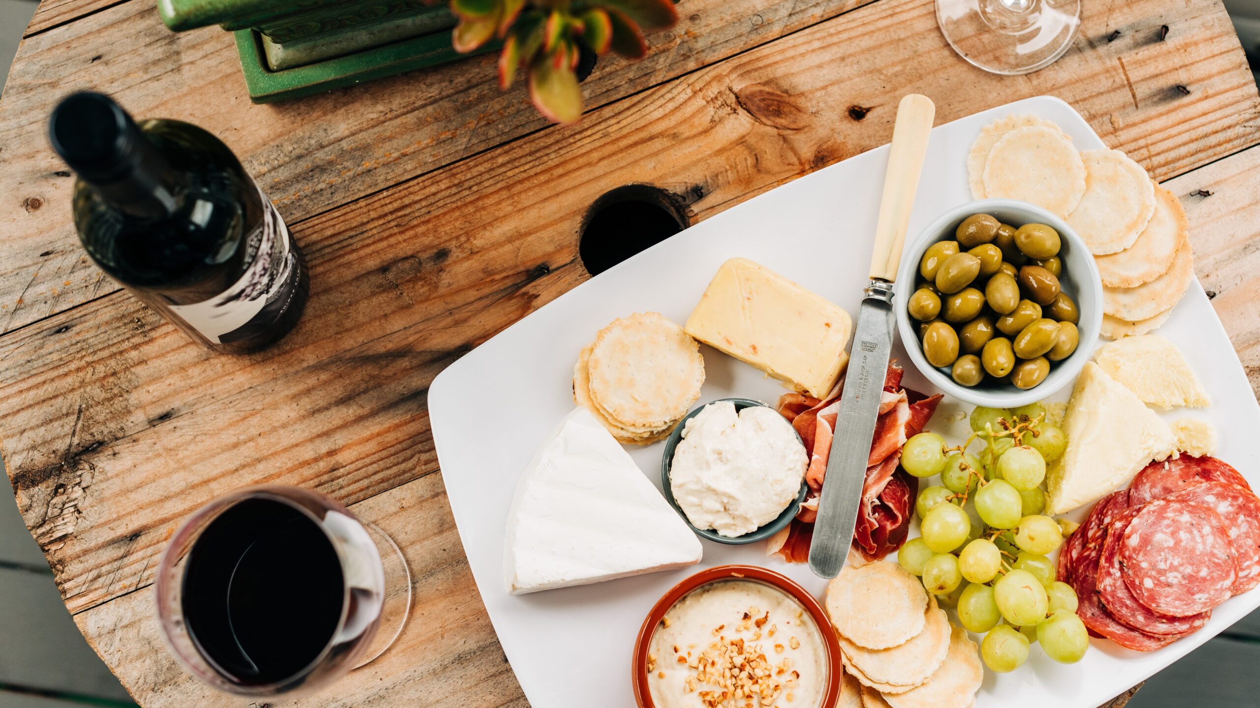 A charcuterie display on a white plate next to two glasses of red wine and a bottle of wine.